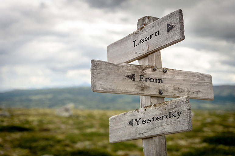 Photograph of a signpost in a rural setting with blurry mountains in the background that has three direction arrows on it with the words Learn From Yesterday separately, each on a different direction arrow.